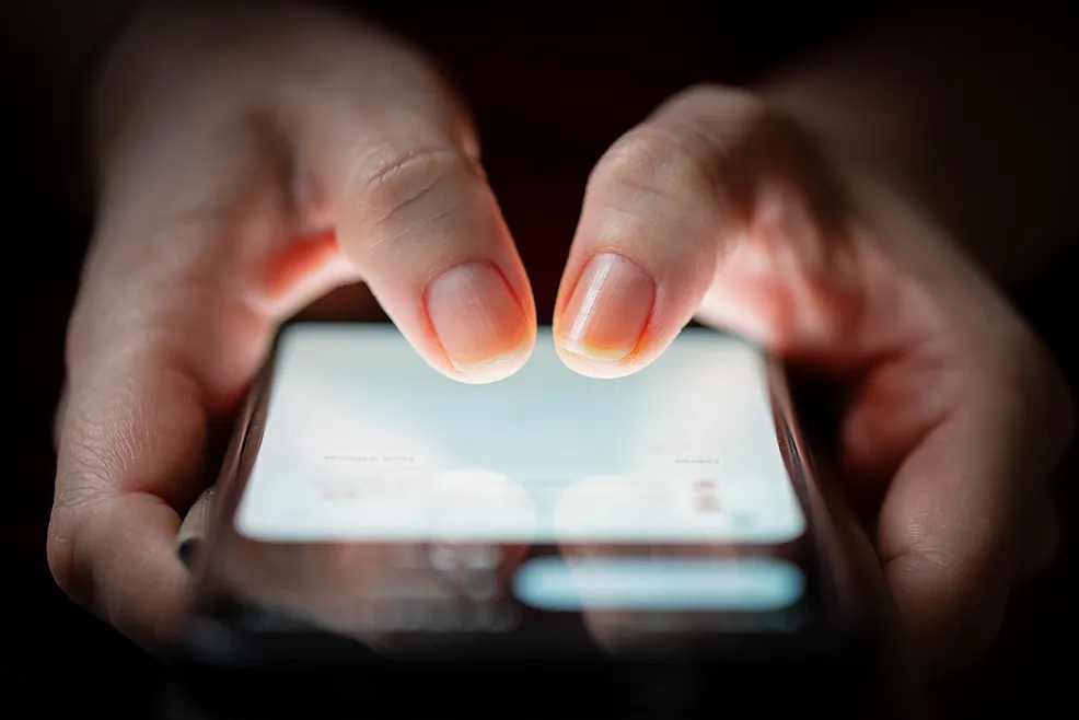Close-up of hands holding and using a smartphone in the dark, with the screen's glow illuminating the fingers, symbolizing contemporary digital dependency and compulsive technology use