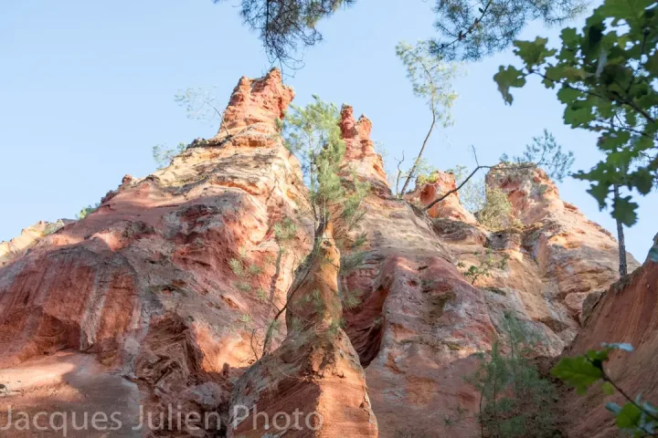 Dramatic red orange ochre rock cliffs with layered sedimentary formations against blue sky with trees